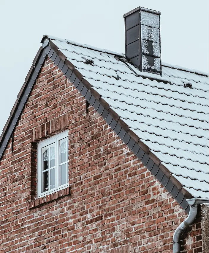 Brick house roof covered with snow and a chimney, with a double window under the roof.