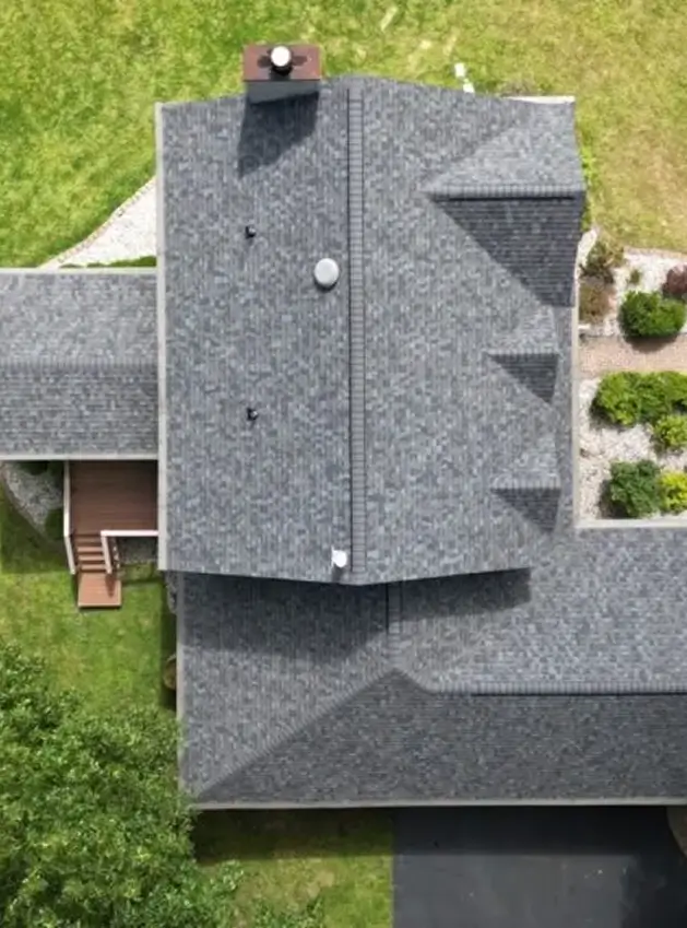 Aerial view of a gray shingled roof on a house with green lawn and shrub landscaping around it.