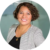 Smiling woman with curly hair wearing a striped blazer and black top against a blurred background.