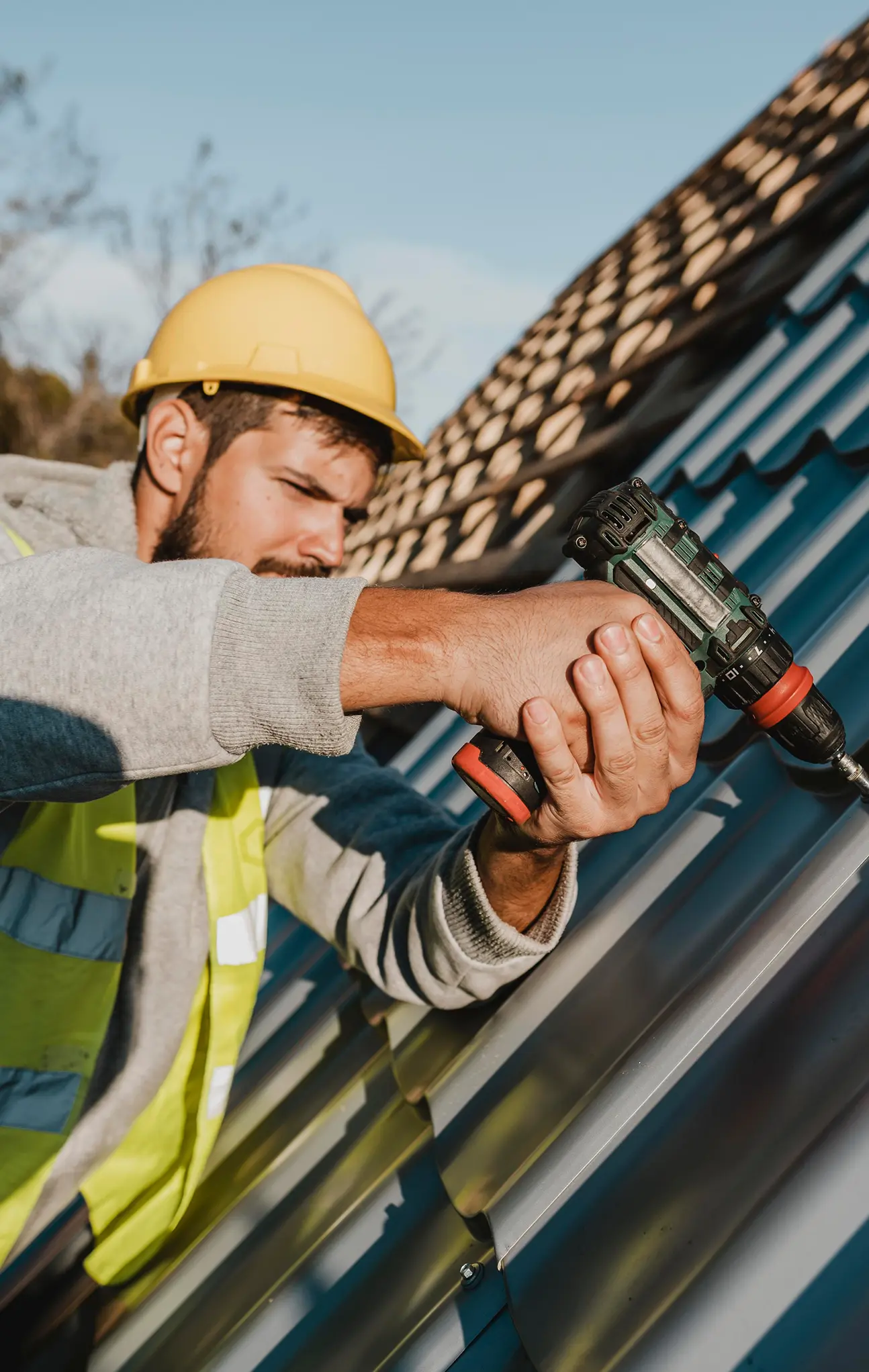 Construction worker wearing yellow hard hat and vest using a drill on metal roofing panels.