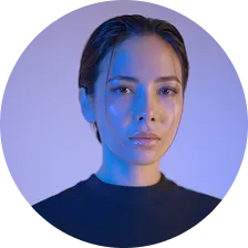 Portrait of a woman with slicked-back hair and blue lighting on her face against a purple background.