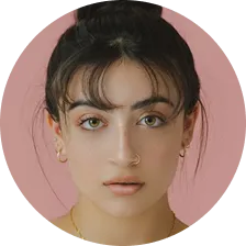 Close-up of a woman with dark hair, hazel eyes, and gold jewelry against a pink background.