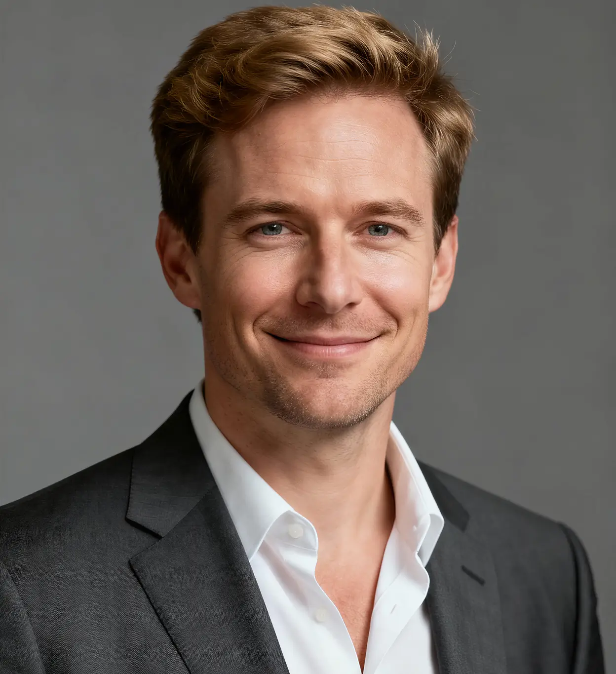 Smiling man with blond hair wearing a white shirt and dark suit jacket against a gray background.