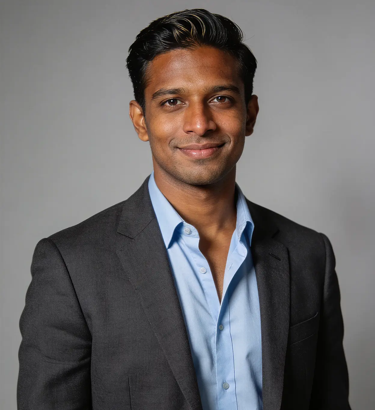 Smiling man wearing a blue shirt and dark blazer against a gray background.