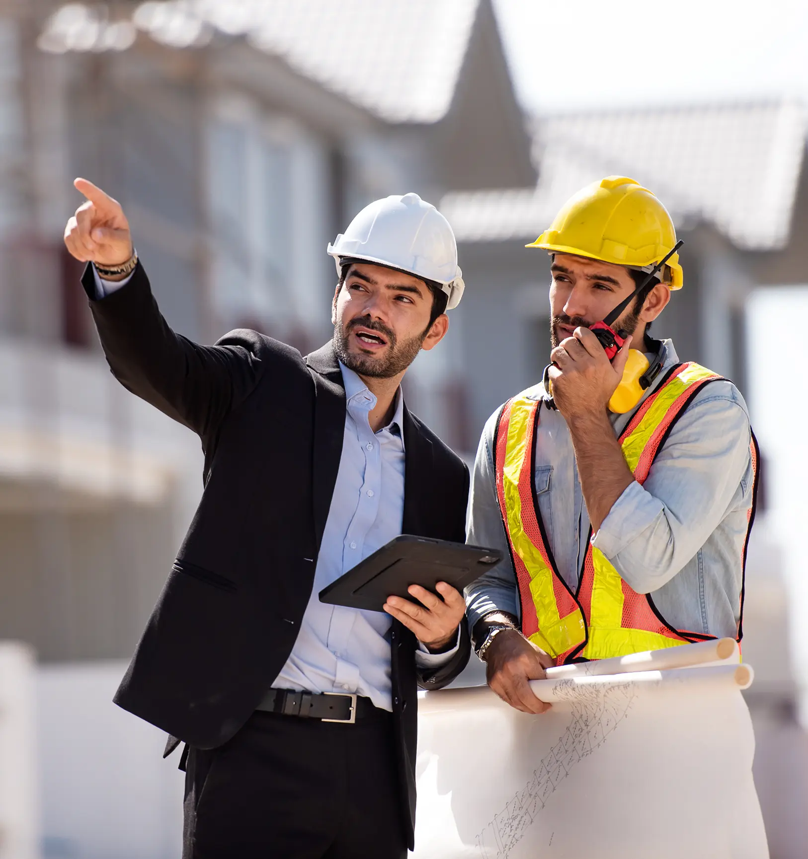Two construction workers wearing safety helmets discuss plans while one points and the other uses a walkie-talkie.