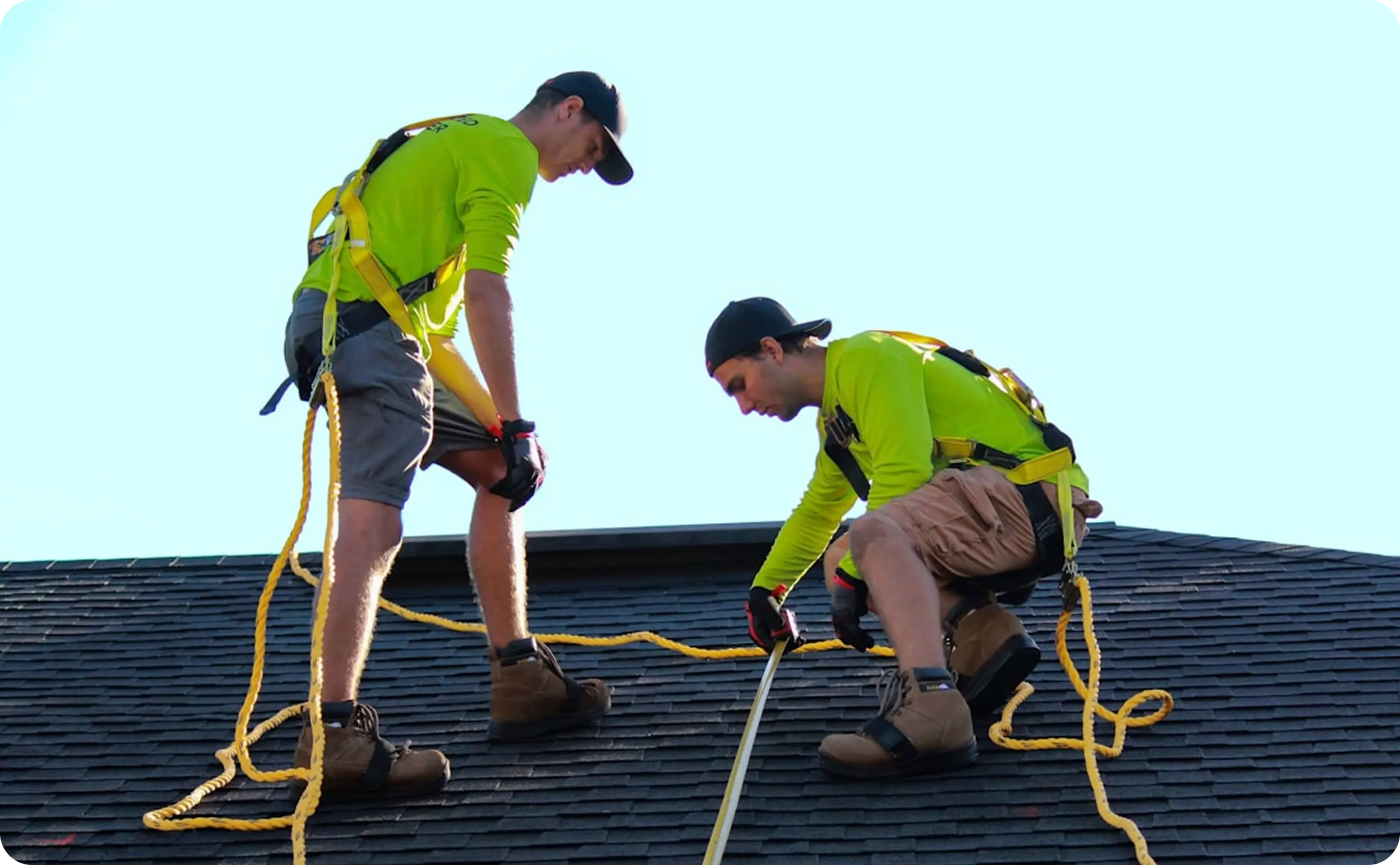 Two workers in safety harnesses measuring a rooftop while wearing boots, gloves, and caps.