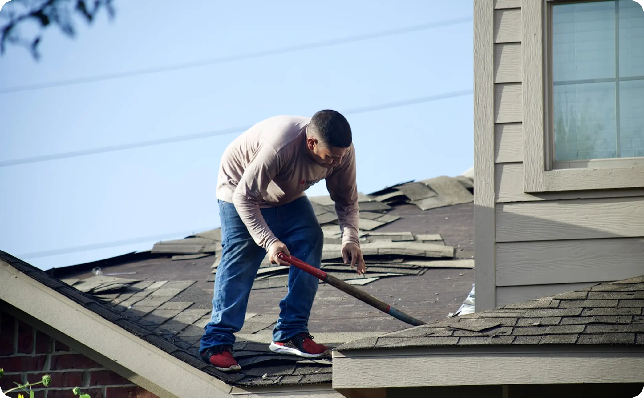 Man on roof removing old shingles with a crowbar during roof repair work.