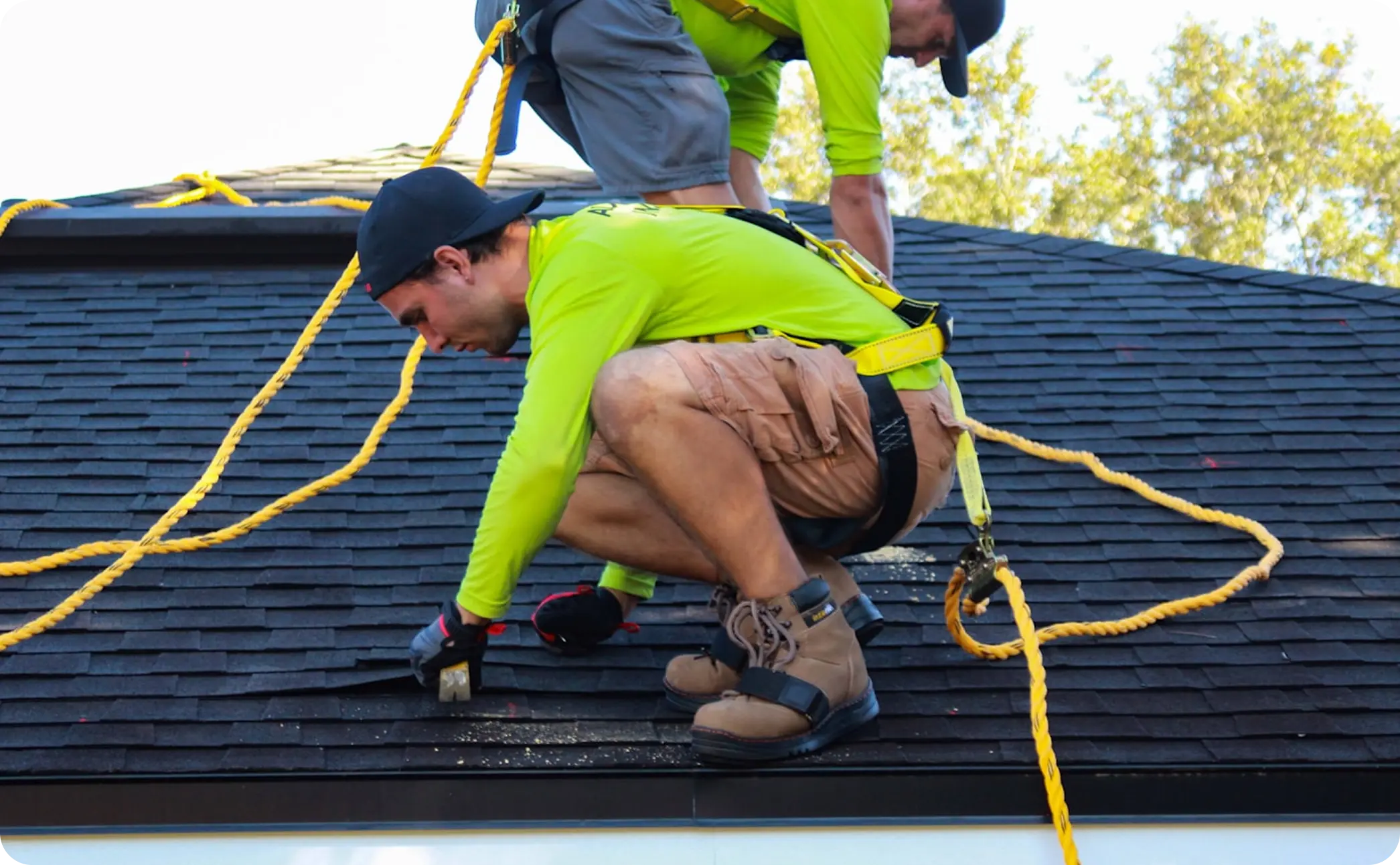 Two workers in safety harnesses working on a roof fixing shingles with safety ropes.