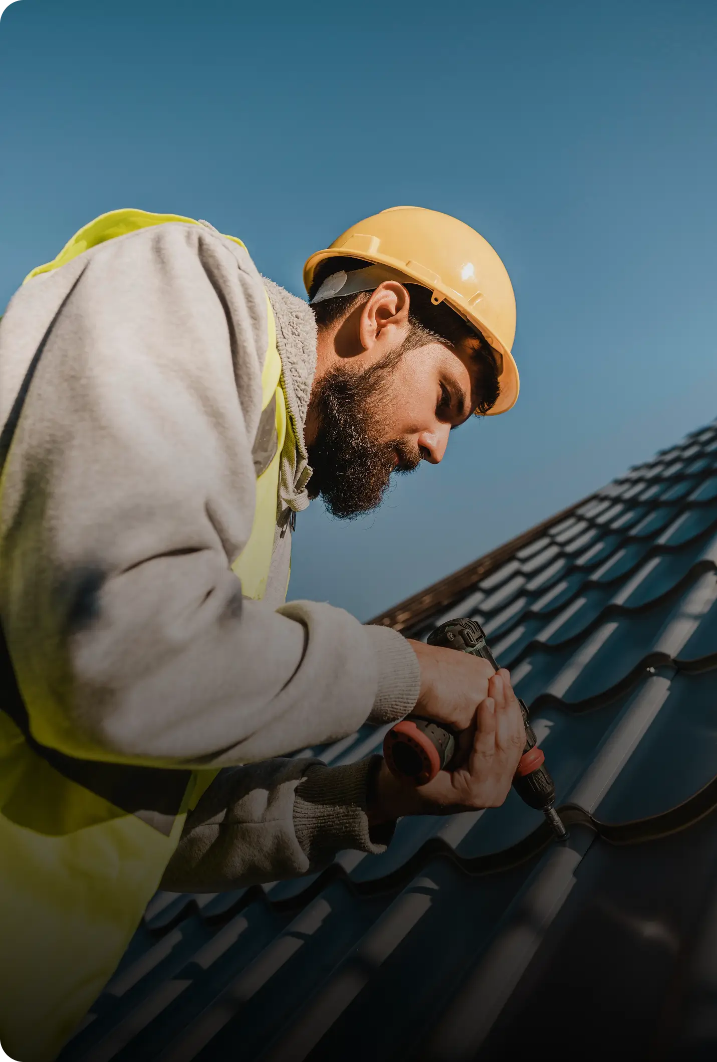 Construction worker in a yellow helmet using a drill on a dark tiled roof under clear sky.