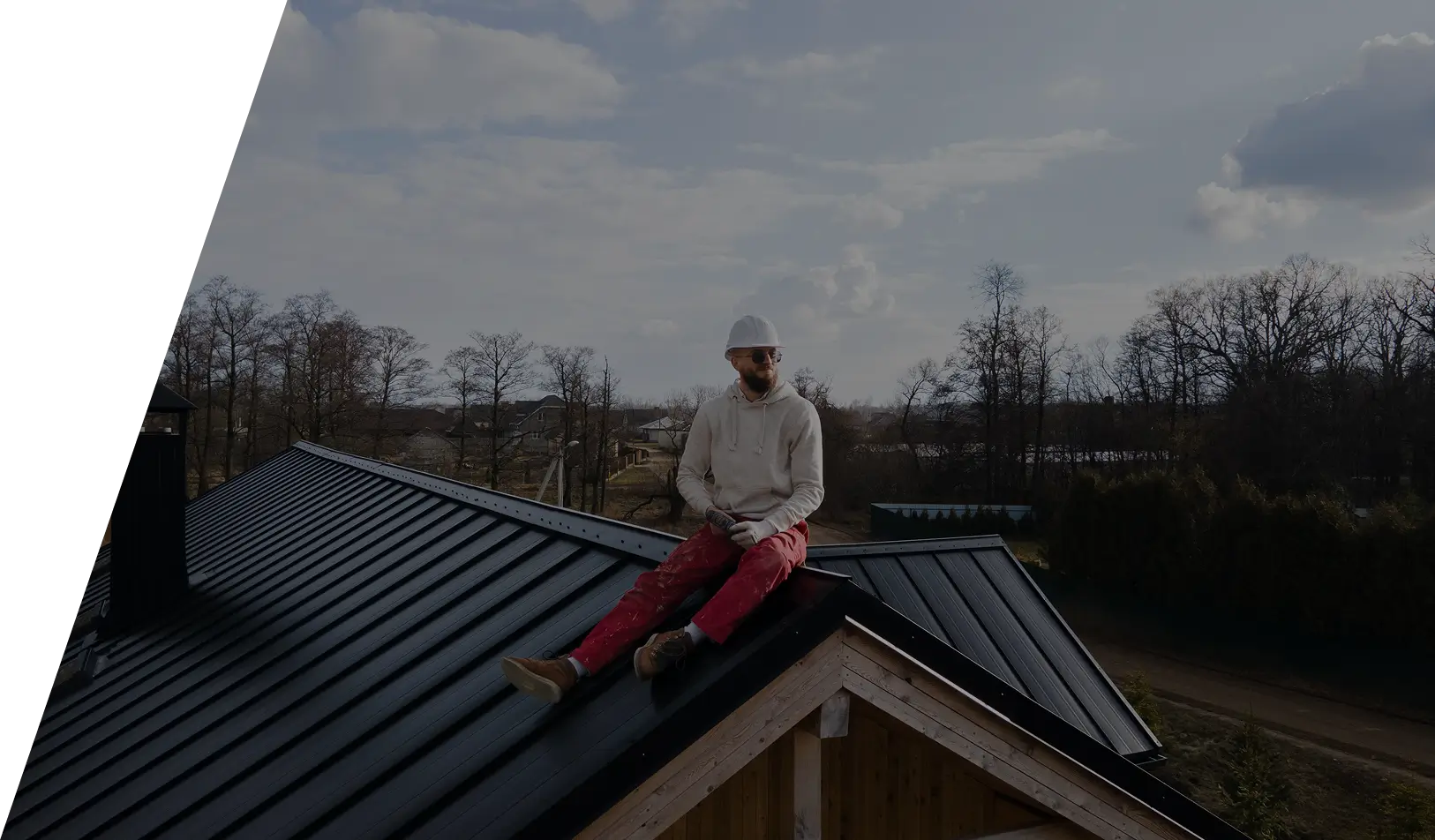 Man in a white helmet and red pants sitting on a metal roof of a wooden house on a cloudy day.