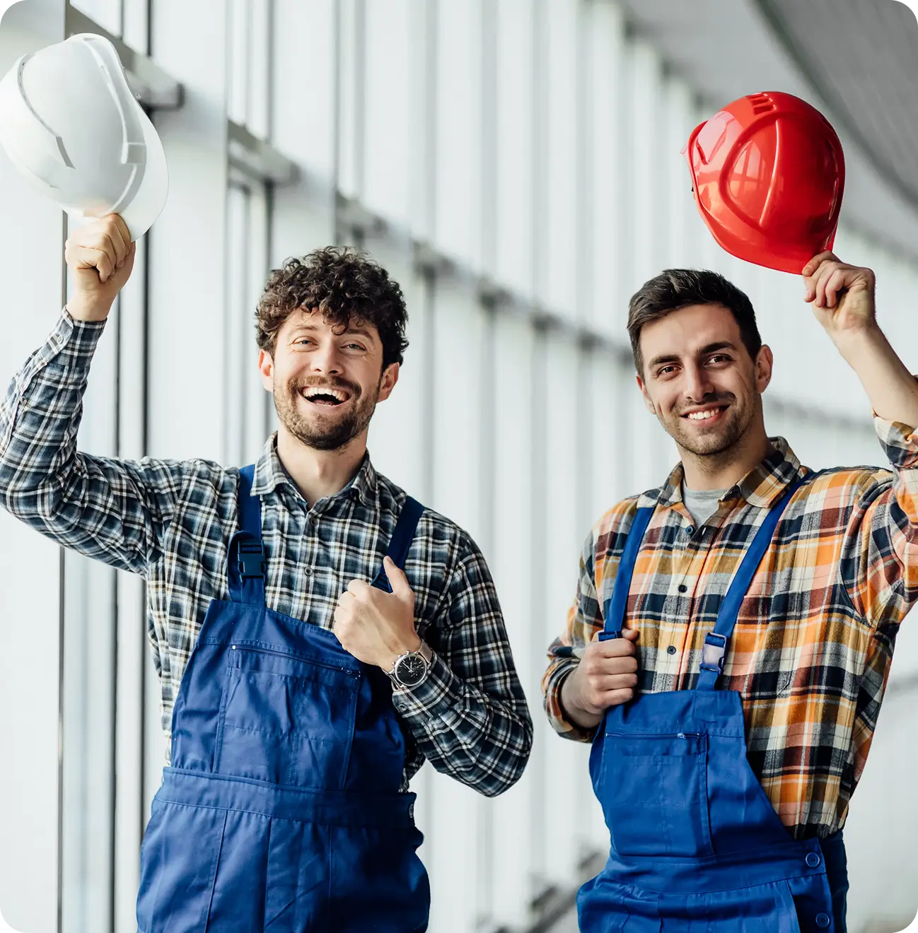 Two male construction workers in blue overalls holding hard hats and smiling inside a bright building.