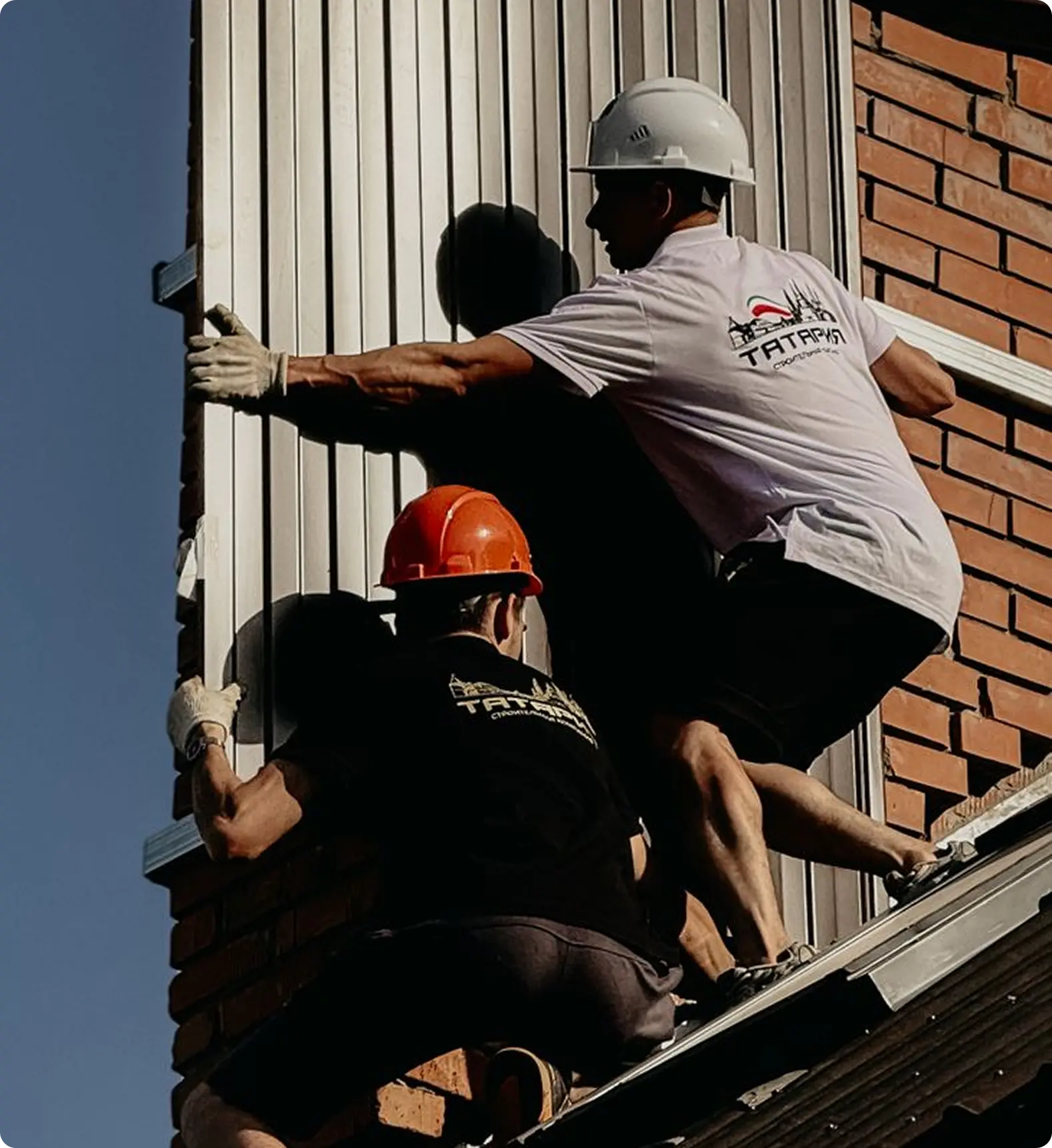 Two construction workers wearing helmets installing a vertical metal panel on a brick wall.